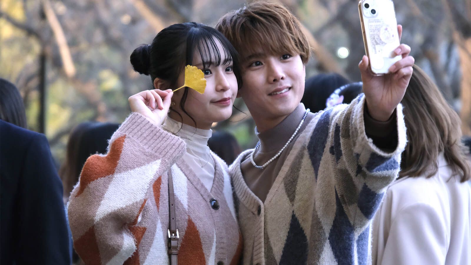 Un couple prend un selfie dans un parc en automne, la femme tient une feuille de ginkgo jaune.