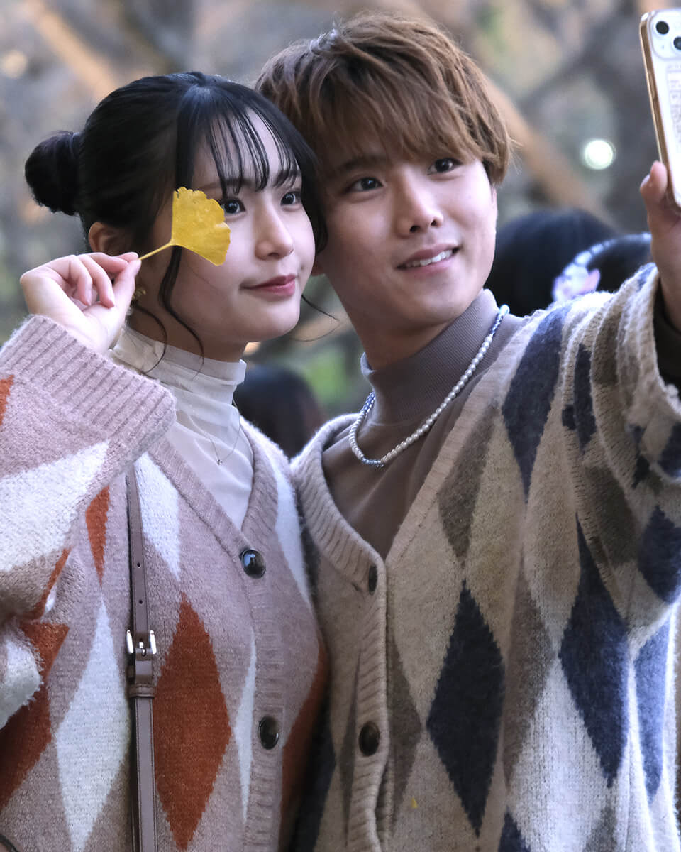Un couple prend un selfie dans un parc en automne, la femme tient une feuille de ginkgo jaune.
