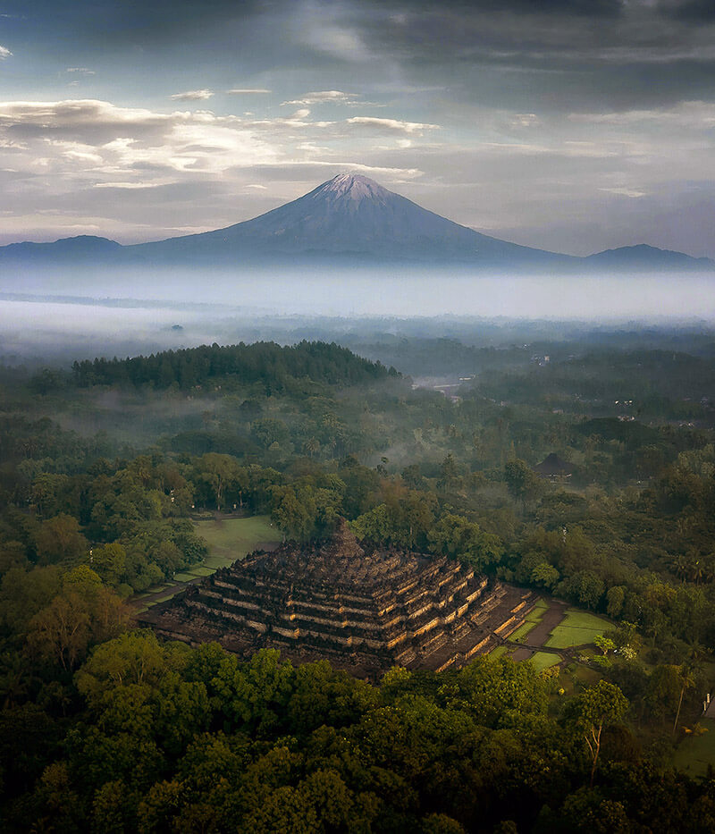 Au premier plan de la photographie, le temple de Borobudur, au second plan, le volcan Merapi, entouré de brume.