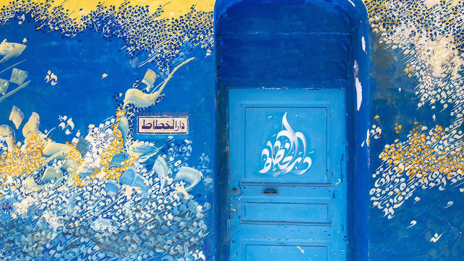 Une fresque murale et une porte bleue ornées de calligraphies dans la médina d'Asilah, au nord du Maroc. L'inscription sur le mur se traduit par « la maison du calligraphe ».
