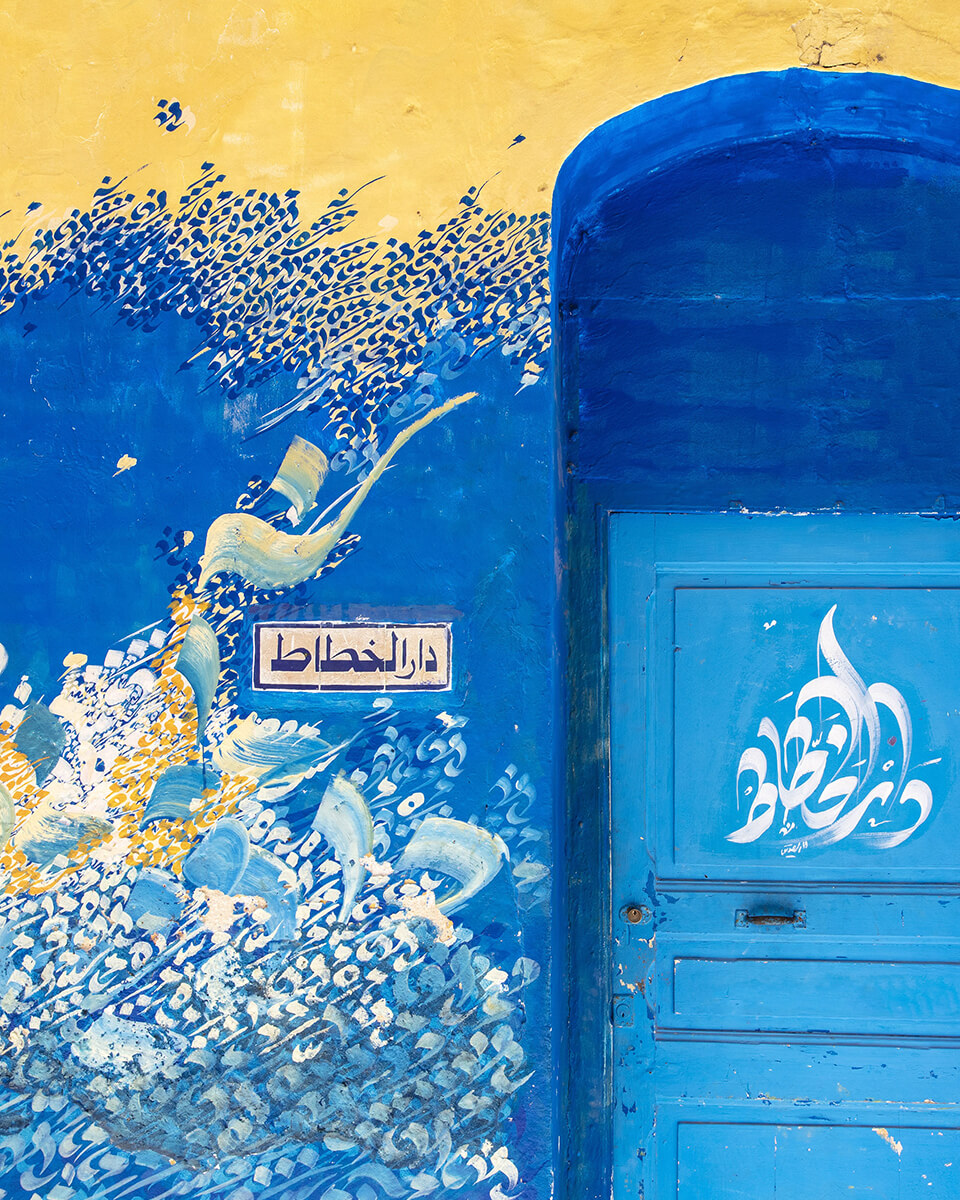 Une fresque murale et une porte bleue ornées de calligraphies dans la médina d'Asilah, au nord du Maroc. L'inscription sur le mur se traduit par « la maison du calligraphe ».