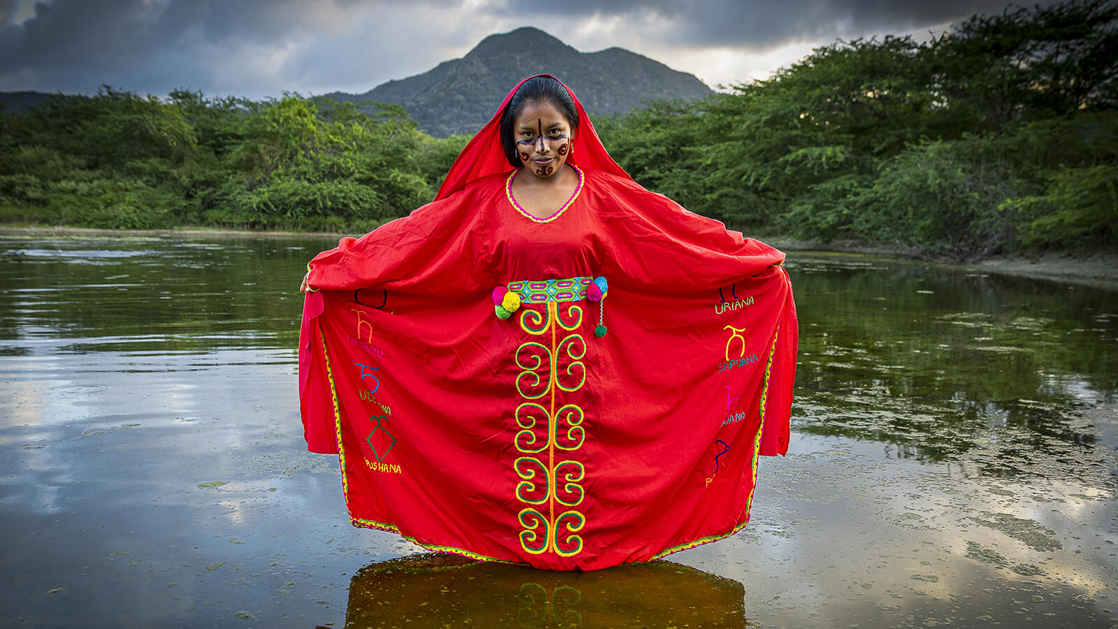 Femme de la communauté Wayuù vêtue de son costume traditionnelle et posant devant un paysage de reliefs verdoyants.