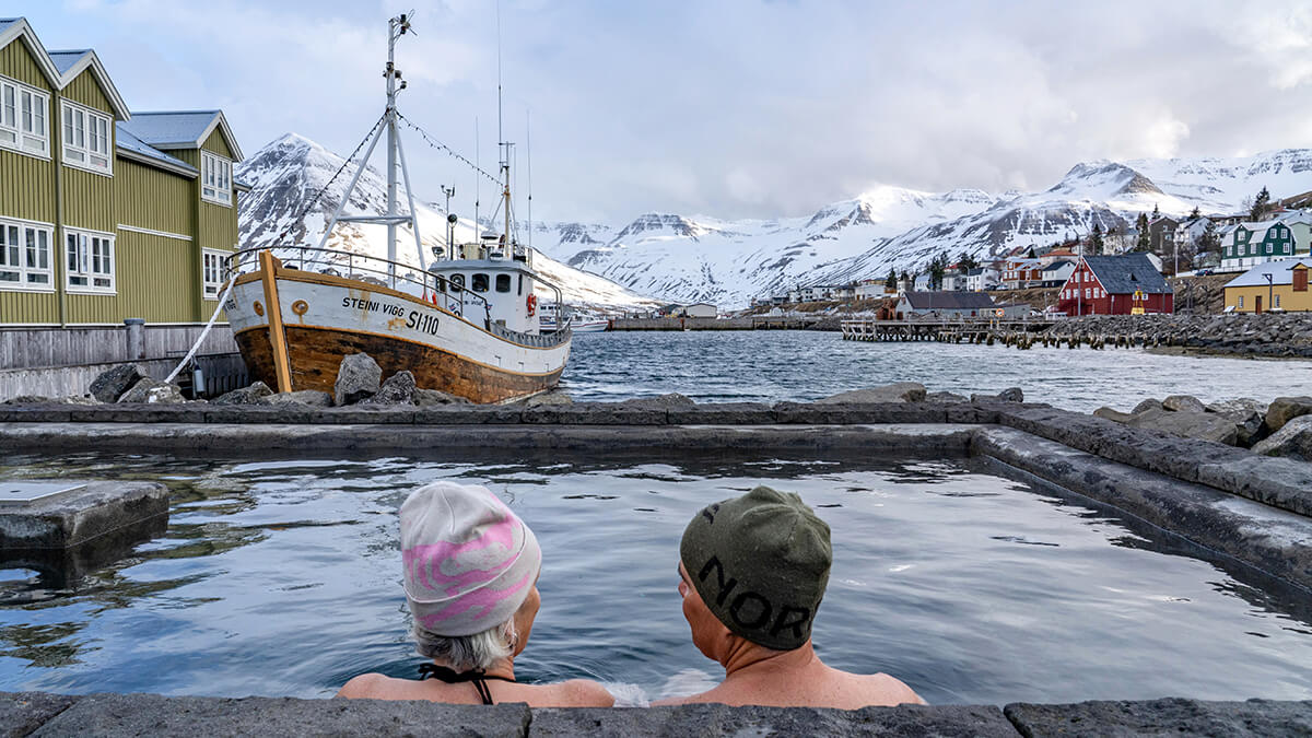 Un couple contemple le petit port et les maisons colorées de de Siglufjörður, un village du nord de l'Islande. Au troisième plan, on aperçoit des sommets enneigés.