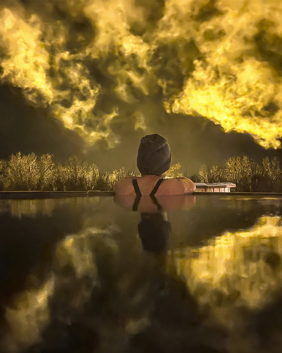 Femme accoudée au rebord d'un bain chaud islandais à l'hôtel Varmaland, près de Borgarnes. La scène se passe de nuit au milieu de nuages, que colorent l'éclairage des bâtiments voisins.